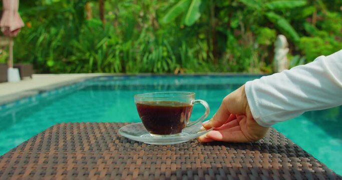 Hot coffee cup on saucer in the garden with green tropical forest and blue swimming pool water. Resort staycation with nature background. Bali Island. Indonesia.