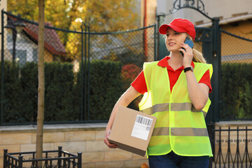 Courier in uniform with parcel talking on smartphone near private house outdoors, space for text