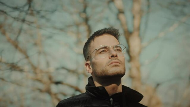 Close-up tracking arc shot revolving around a man's face. A young depressed, sad, and unhappy Caucasian male stands out in nature in a forest with trees. Suffering from mental health. Slow motion.
