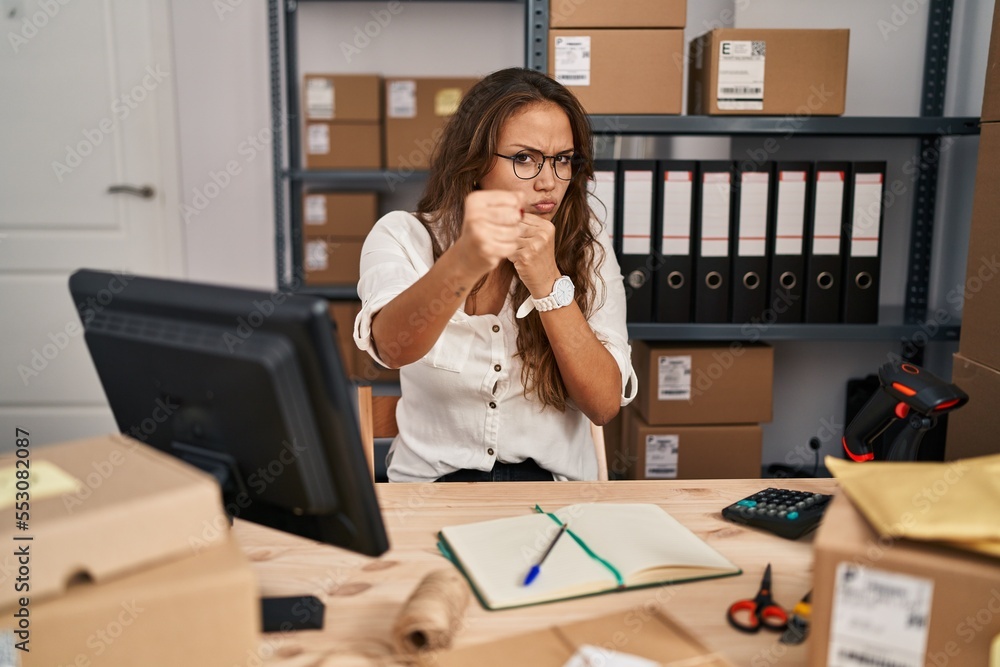 Wall mural Young hispanic woman working at small business ecommerce ready to fight with fist defense gesture, angry and upset face, afraid of problem