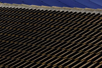 Gray colored cement roof tiles lit with side lighting from the setting sun. Old house roofing, gray concrete tiles showing years of exposure to the elements, with modern solar panels in the background