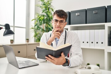 Young hispanic man doctor using laptop reading book at clinic