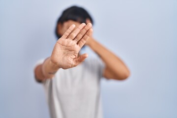 Young hispanic woman wearing casual t shirt over blue background covering eyes with hands and doing...