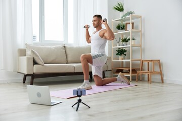 Obraz premium Man sports, watching a workout tape on his phone and repeating exercises sports blogger with dumbbells, pumped up man fitness trainer works out at home, the concept of health and body beauty