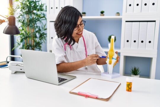 Young latin woman wearing doctor uniform holding anatomical model of knee at clinic
