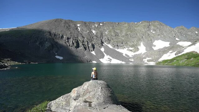 Lonely Woman Sitting on Rock in Front of Idyllic Alpine Lake Under Mountain Hills on Sunny Summer Day