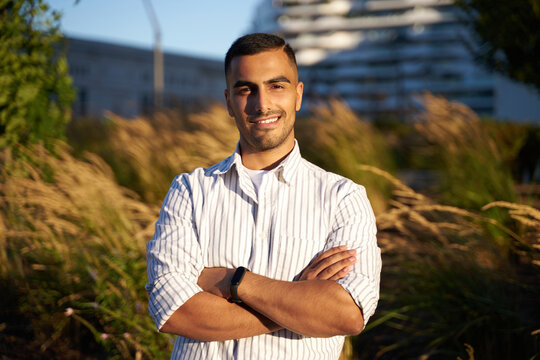Young smiling middle eastern man with arm crossed  wearing stylish shirt looking at camera standing in park 