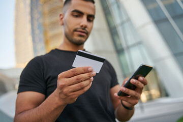 Handsome Middle Eastern man holding credit card and mobile phone shopping online on the street, selective focus. Modern technology concept 