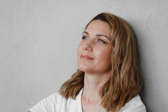 Closeup portrait of happy creative young woman smiling and looking directly to the camera over grey wall. Female at her 40s relaxed in white t-shirt