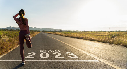 Fototapeta New year 2023 or start straight concept.word 2023 written on the asphalt road and athlete woman runner stretching leg preparing for new year at sunset.Concept of challenge or career path and change. obraz