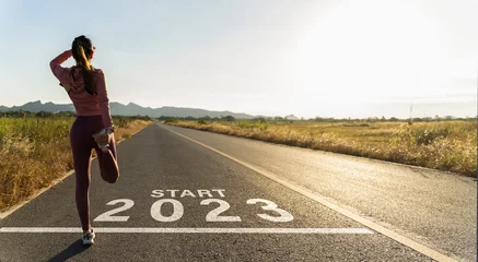 Zelfklevend Fotobehang New year 2023 or start straight concept.word 2023 written on the asphalt road and athlete woman runner stretching leg preparing for new year at sunset.Concept of challenge or career path and change. © Yingyaipumi