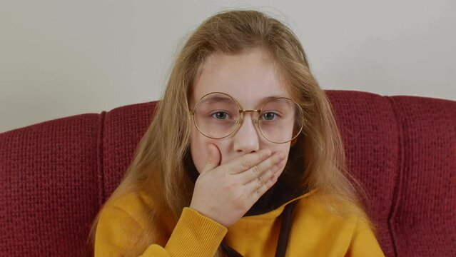 Funny little girl with long blond hair wearing round big glasses and light mustard sweatshirt. Girl seriously looks at camera, then smiles showing toothless mouth and funny covers it with her palm