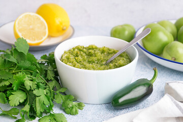 Bowl of tasty green salsa sauce, fresh jalapeno pepper and cilantro on light table