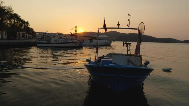 Closeup of a blue fishing paddle boat tied to the pier swaying on the waves. Ved in front. In the background pleasure boats, mountains and sunset. People walk along the waterfront