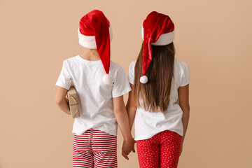 Little children in pajamas with Christmas gift holding hands near beige wall, back view