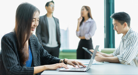 young businesswoman sitting, smiling, and working with laptop. man works with tablet at background. young man and woman standing discussion at window background in relaxing workplace