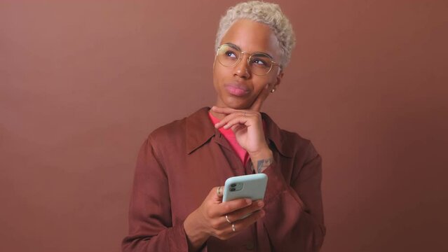 Young puzzled African American woman with mobile phone in hand touches chin thinks about writing resume in smartphone application to search for new job stands posing in burgundy studio