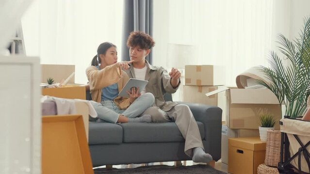 Young happy Asia couple sit on sofa couch use digital tablet online shopping furniture from decoration store in living room at home. Husband wife move new house, Apartment Rent And Ownership Concept.