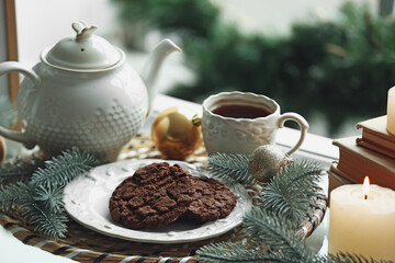 Plate with cookies, teapot, cup of tea, Christmas branches and balls on windowsill, closeup