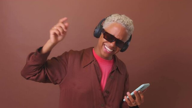 Young joyful African American woman in headphones and sunglasses holding phone and listening to music having fun at solidarity party and dancing stands on plain burgundy background. Disco, nightclub