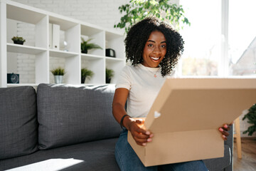 Young african woman sit on couch at home unpacking parcel cardboard box with online purchase