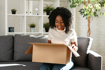Young african woman sit on couch at home unpacking parcel cardboard box with online purchase - obrazy, fototapety, plakaty