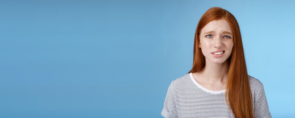 Displeased young awkward redhead girl cringe full disbelief smirking frowning confused look questioned doubtful hearing nuisance dumb story standing blue background uncertain - obrazy, fototapety, plakaty
