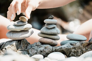 Woman with daughter bilds stones pyramid on seashore on a sunny day on the blue sea background. Happy family holidays. Pebble beach, calm sea. Concept of happy vacation on the sea, meditation, spa