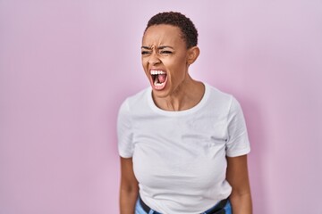 Beautiful african american woman standing over pink background angry and mad screaming frustrated...