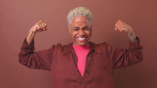 Young confident strong African American woman laughing and showing muscles rejoicing at achievements in sports or winning competitions in fitness club stands in strongman pose in burgundy studio
