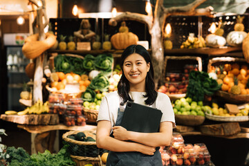 Smiling entrepreneur holding a digital tablet standing at an outdoor market. Asian woman in an apron looking at camera while standing against a street food market. - obrazy, fototapety, plakaty