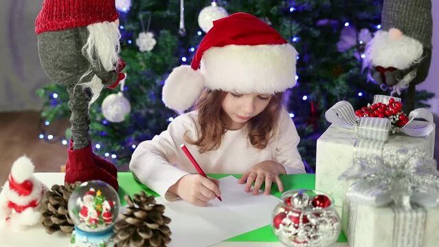 A little girl in a red Santa's Hat writes a letter to Santa Claus with her wishes under the Christmas tree. Christmas and New Year atmosphere. Slow motion