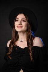 Beautiful young woman in black clothes with a black hat. Photo in the studio on a dark background with blue backlight.