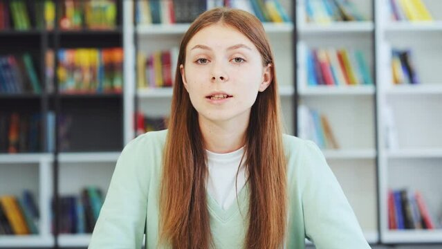 Teen girl waving hand talking looking at camera in library. Caucasian teenager recording vlog, streaming for social media, video calling in online chat. Web cam view headshot. Distance learning