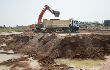 At the construction site, large working machines - an orange crawler excavator and a gray dump truck in the process of working - loading soil from pits flooded with groundwater