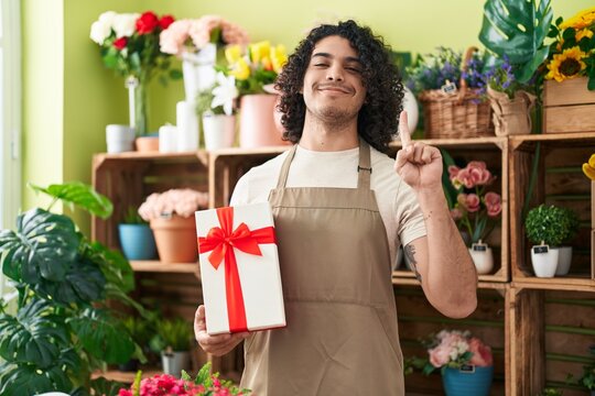 Hispanic man with curly hair working at florist shop holding gift smiling with an idea or question pointing finger with happy face, number one