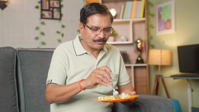 Middle aged man eating fruit salad while sitting on sofa at home - concept of health lifestyle, dieting and wellbeing