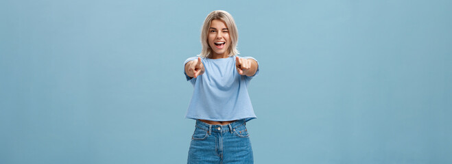 Happy enthusiastic stylish blonde female with tattoos pointing at camera as if picking or making hint smiling joyfully and winking from amazement and happiness posing over blue background - obrazy, fototapety, plakaty