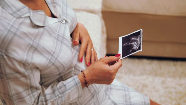 Beautiful pregnant woman holding and looking ultrasound photo of her newborn baby on the bed in bedroom. Maternity expecting healthcare concept. Future mother stroking belly with unborn baby 