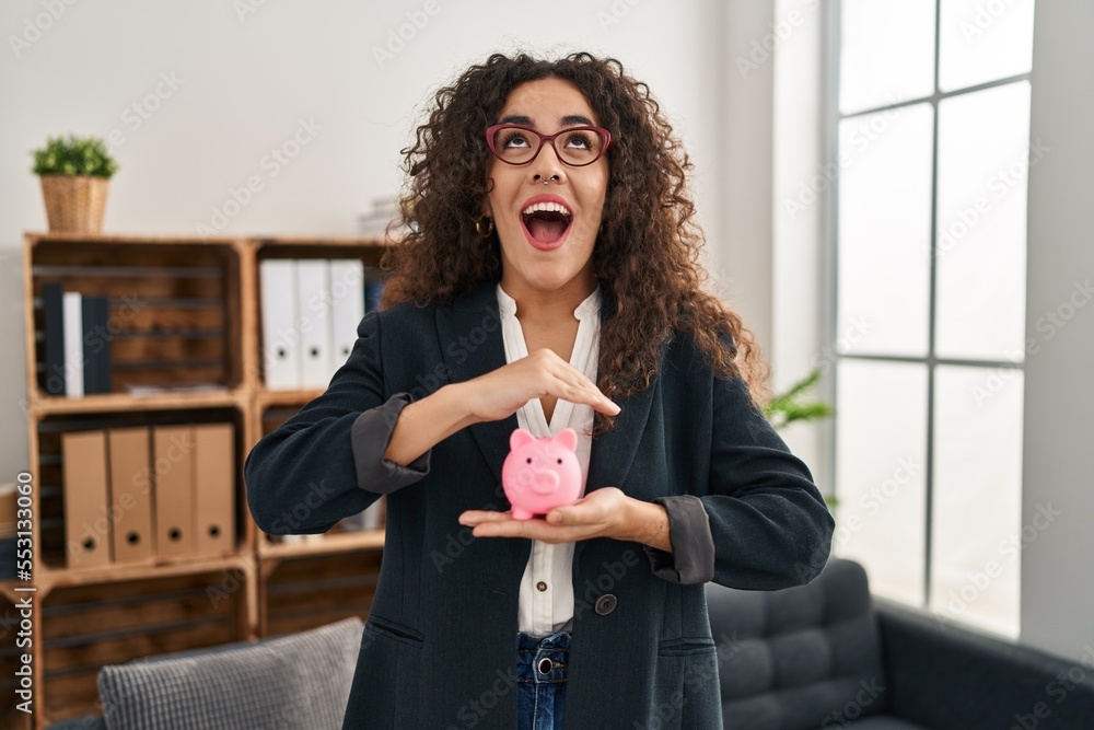 Poster Young hispanic woman holding piggy bank angry and mad screaming frustrated and furious, shouting with anger looking up.