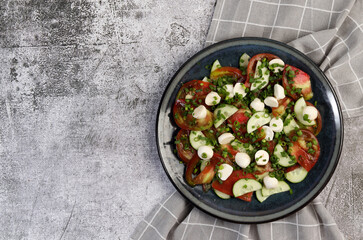 Summer homemade Tomato, mozzarella and cucumbers salad on a round plate on a dark gray background. Top view, flat lay