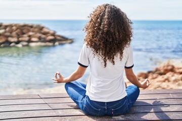 Young hispanic woman sitting on bench doing yoga exercise at seaside