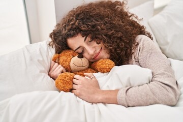 Young hispanic woman hugging teddy bear lying on bed sleeping at bedroom