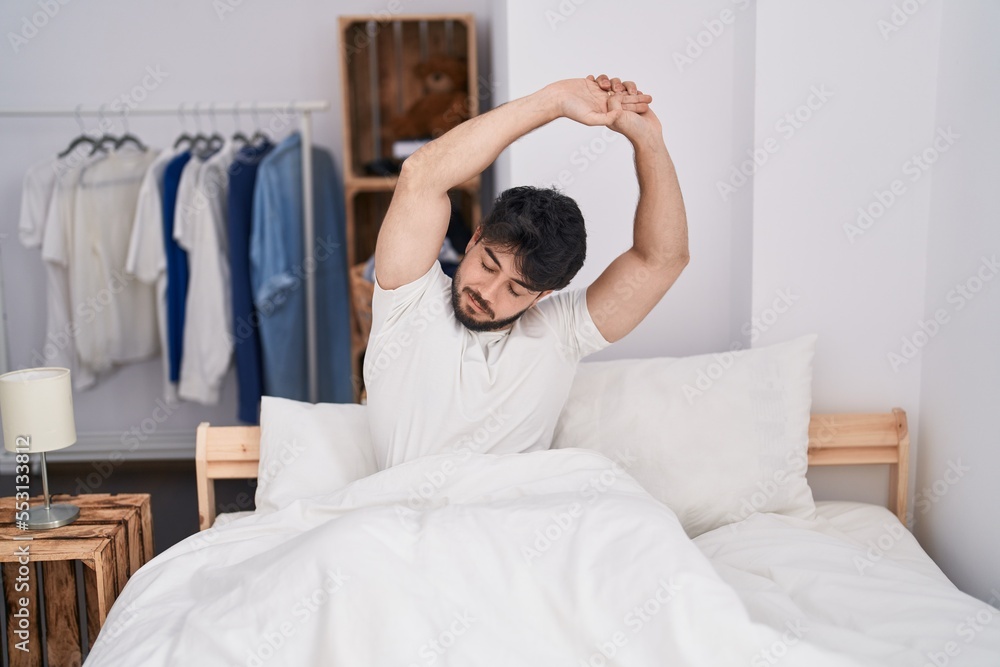 Sticker young hispanic man waking up stretching arms at bedroom