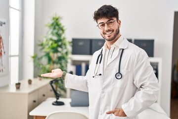 Young hispanic man wearing doctor uniform doing welcome gesture with hand at clinic