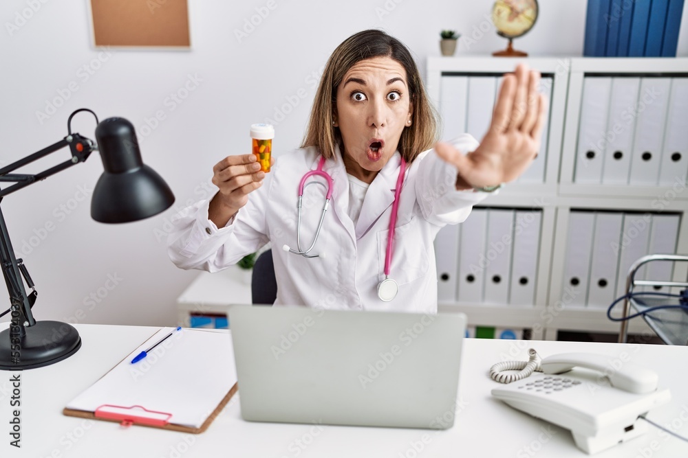 Canvas Prints young hispanic woman wearing doctor uniform holding pills at the clinic doing stop gesture with hand