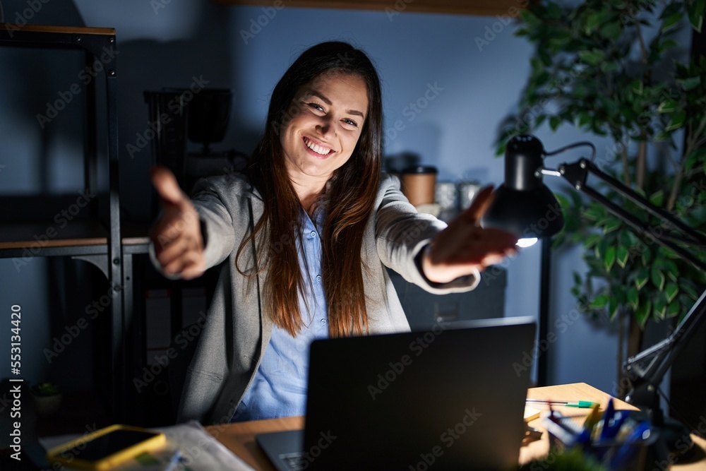 Wall mural Young brunette woman working at the office at night looking at the camera smiling with open arms for hug. cheerful expression embracing happiness.