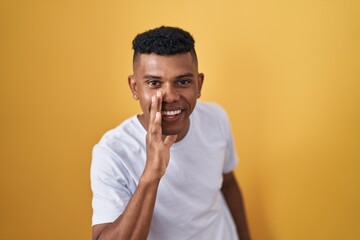 Young hispanic man standing over yellow background hand on mouth telling secret rumor, whispering malicious talk conversation