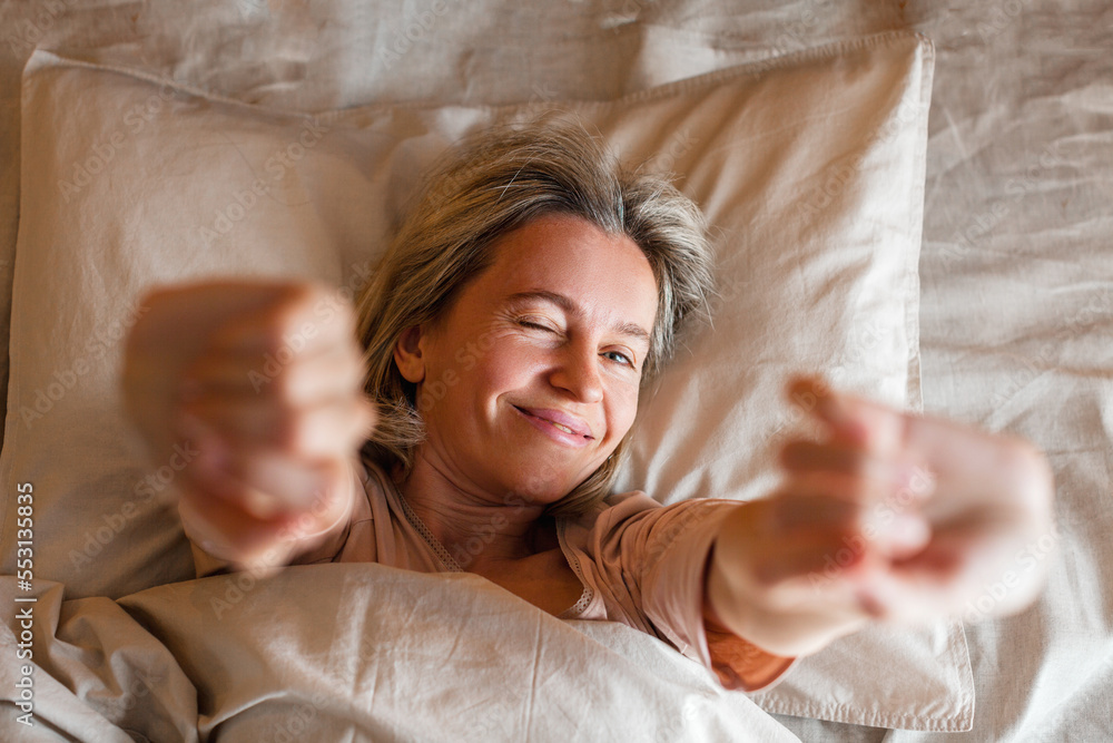 Wall mural Happy middle aged woman sits on bed and stretching arms