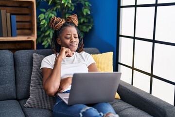 African woman with braided hair using laptop at home thinking concentrated about doubt with finger...